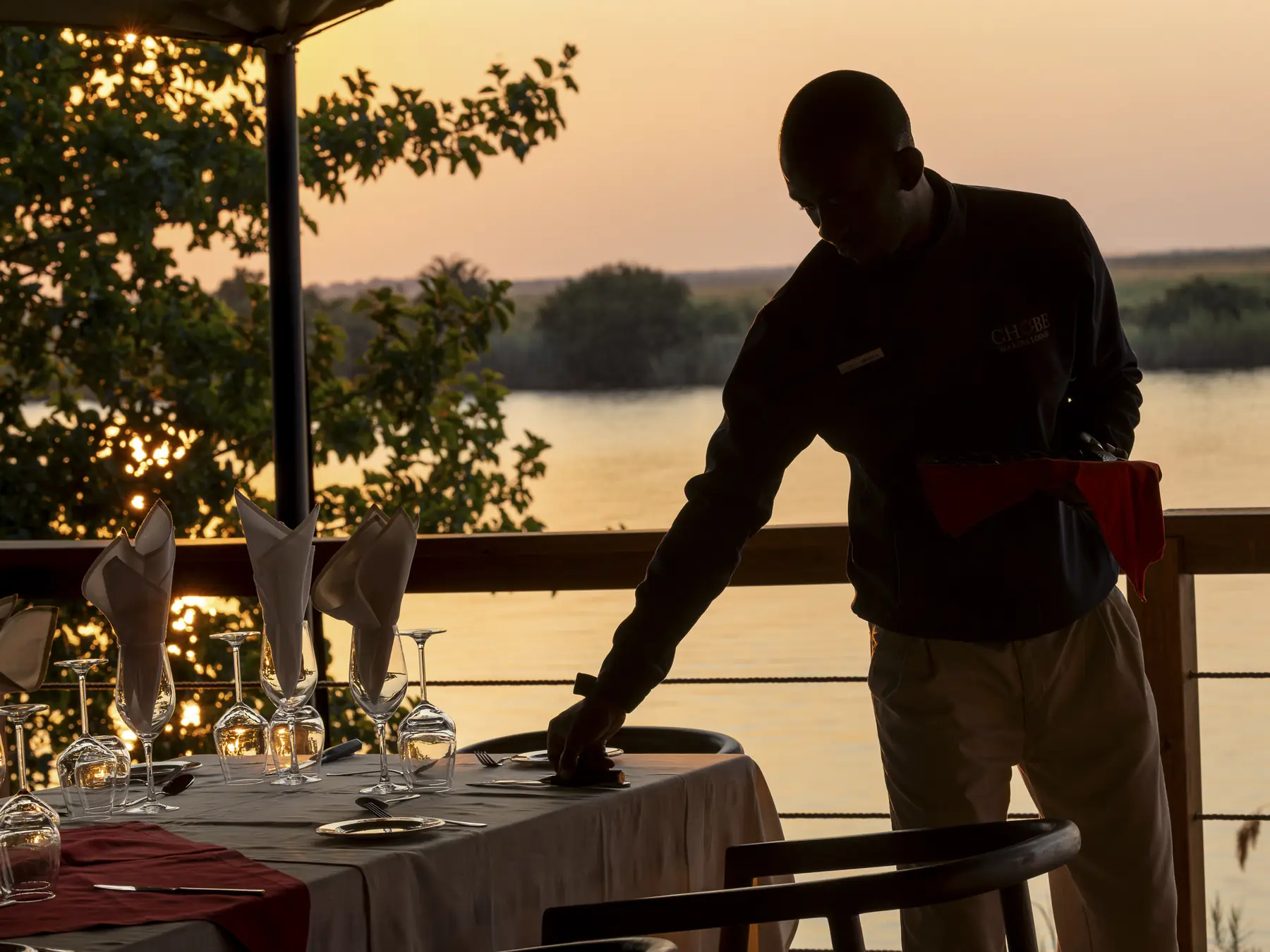 A waiter sets a table on the river deck as the sun goes down over the Chobe
